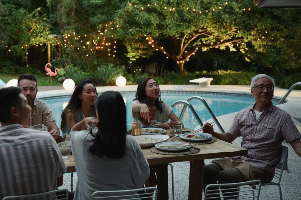 A family sits around an outdoor dining table beside a pool at dusk, with twinkling lights in the trees beyond
