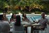 A family sits around an outdoor dining table beside a pool at dusk, with twinkling lights in the trees beyond