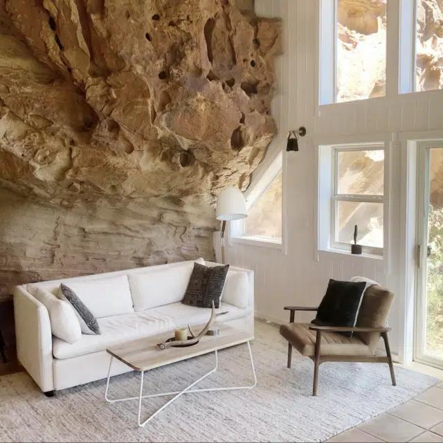 The living room of Cliff House, Colorado, furnished by a white sofa and coffee table and brown armchair, with the sandstone of Sage Canyon forming one wall of the space