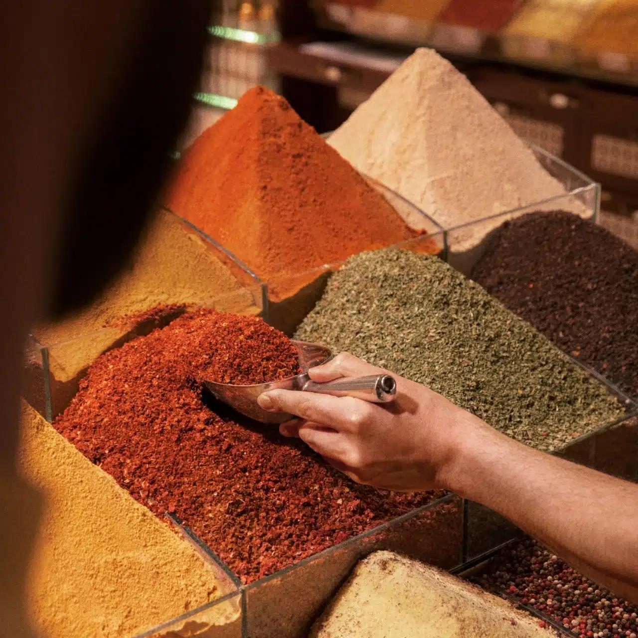 A hand scooping spices from brightly colored piles of powder at a market stall