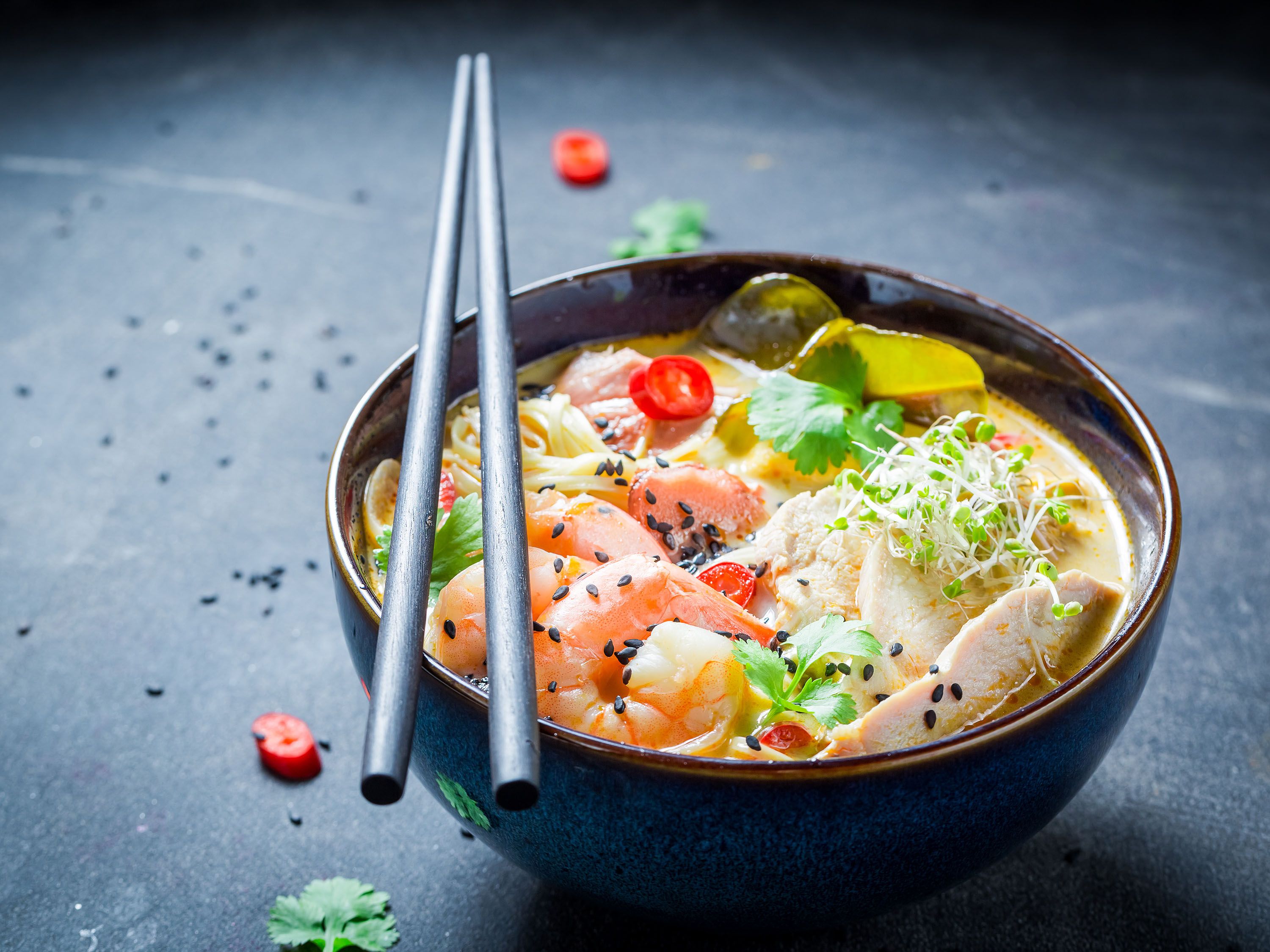 Chopsticks rest on a bowl filled with broth, shrimp, vegetables and sesame seeds.