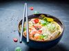 Chopsticks rest on a bowl filled with broth, shrimp, vegetables and sesame seeds.