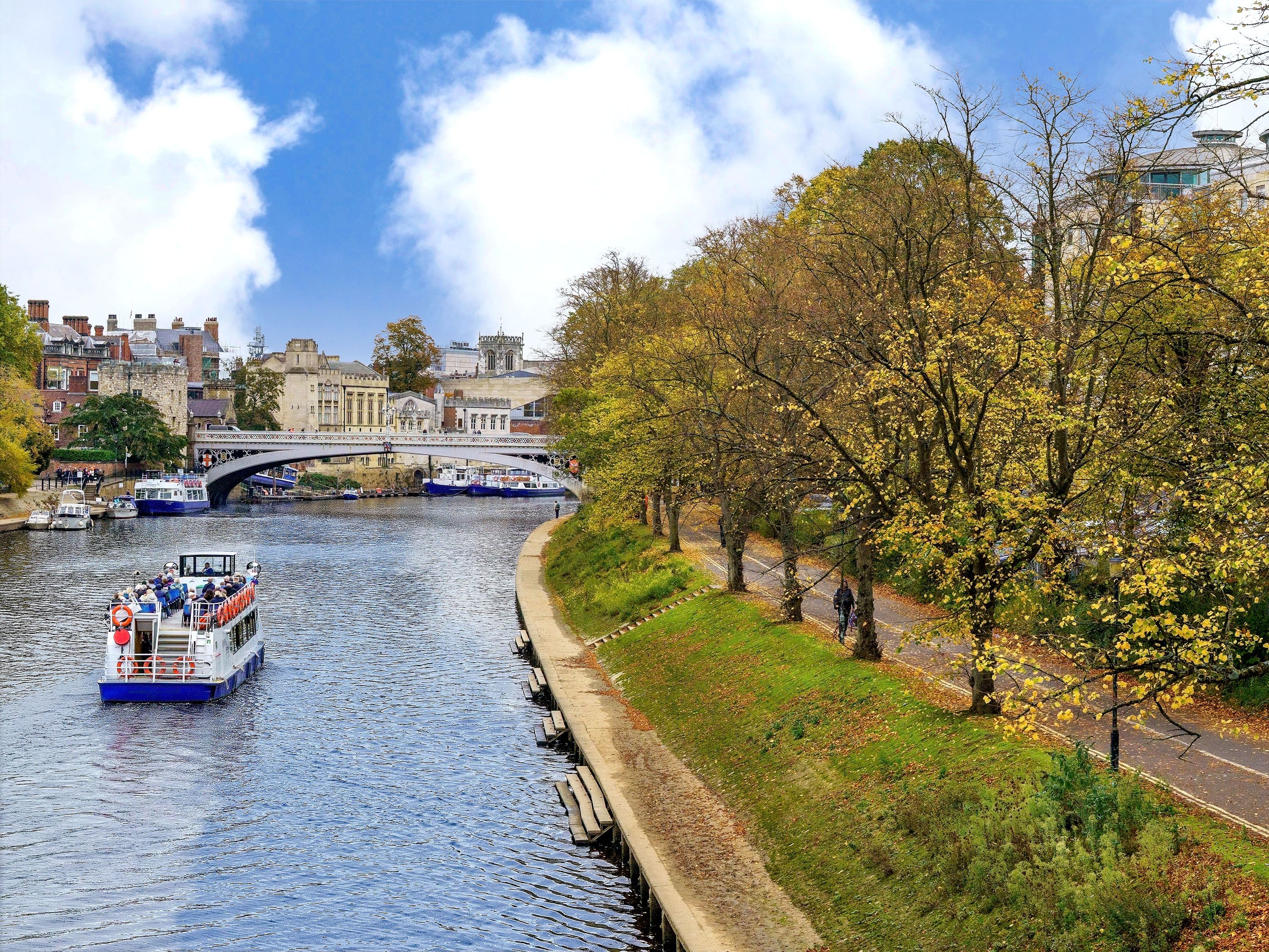 River boat heading toward bridge with walking and bicycle path along the river's edge.