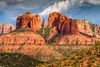 Red and tan colored striated buttes with low desert scrub under cloudy sky.