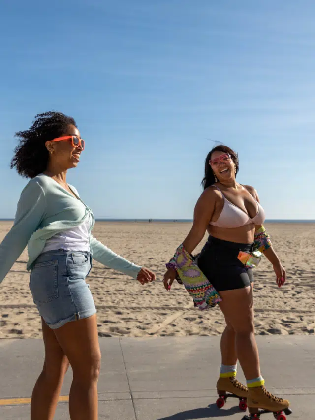 Two women rollerskate in the sunshine beside a beach in Los Angeles, California