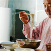 A man lifts noodles from a metal bowl with chopsticks, surrounded by smaller ceramic bowls