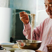 A man lifts noodles from a metal bowl with chopsticks, surrounded by smaller ceramic bowls
