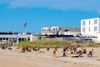 People sunbathing and lounging on a sandy beach with grassy dunes, nearby buildings, and an American flag flying under a clear blue sky.