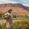 A blonde haired woman wearing a long beige skirt and white shirt stands in front of a red mountain range carrying a large basket