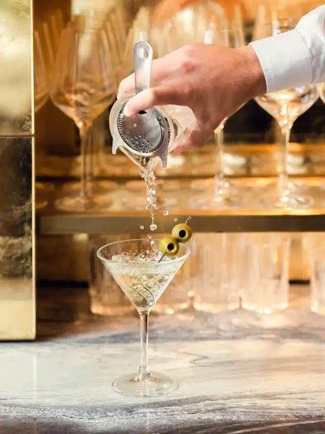 A mixologist's hand pours a martini from a shaker into a glass with two olives on a stick