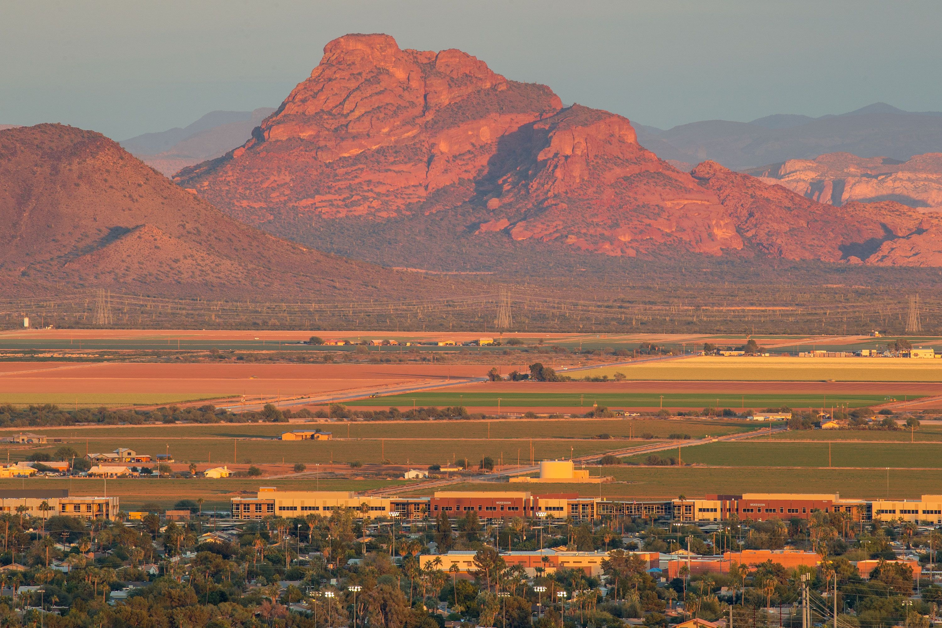 A wide landscape view of a red rock mountain glowing at sunset with fields, houses, and low-rise buildings in the foreground.