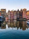Eight crooked canalhouses, known as the Dancing Houses of Amsterdam, are seen across the canal on a sunny day