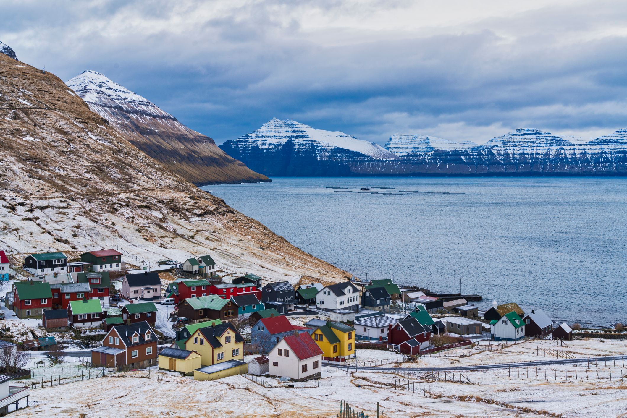 Red, yellow and green houses sit between a snow-dusted hillside and the fjord in the Faroe Islands, with layered peaks across the water.