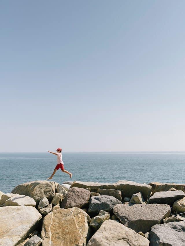 A child jumps along giant rocks with the sea beyond