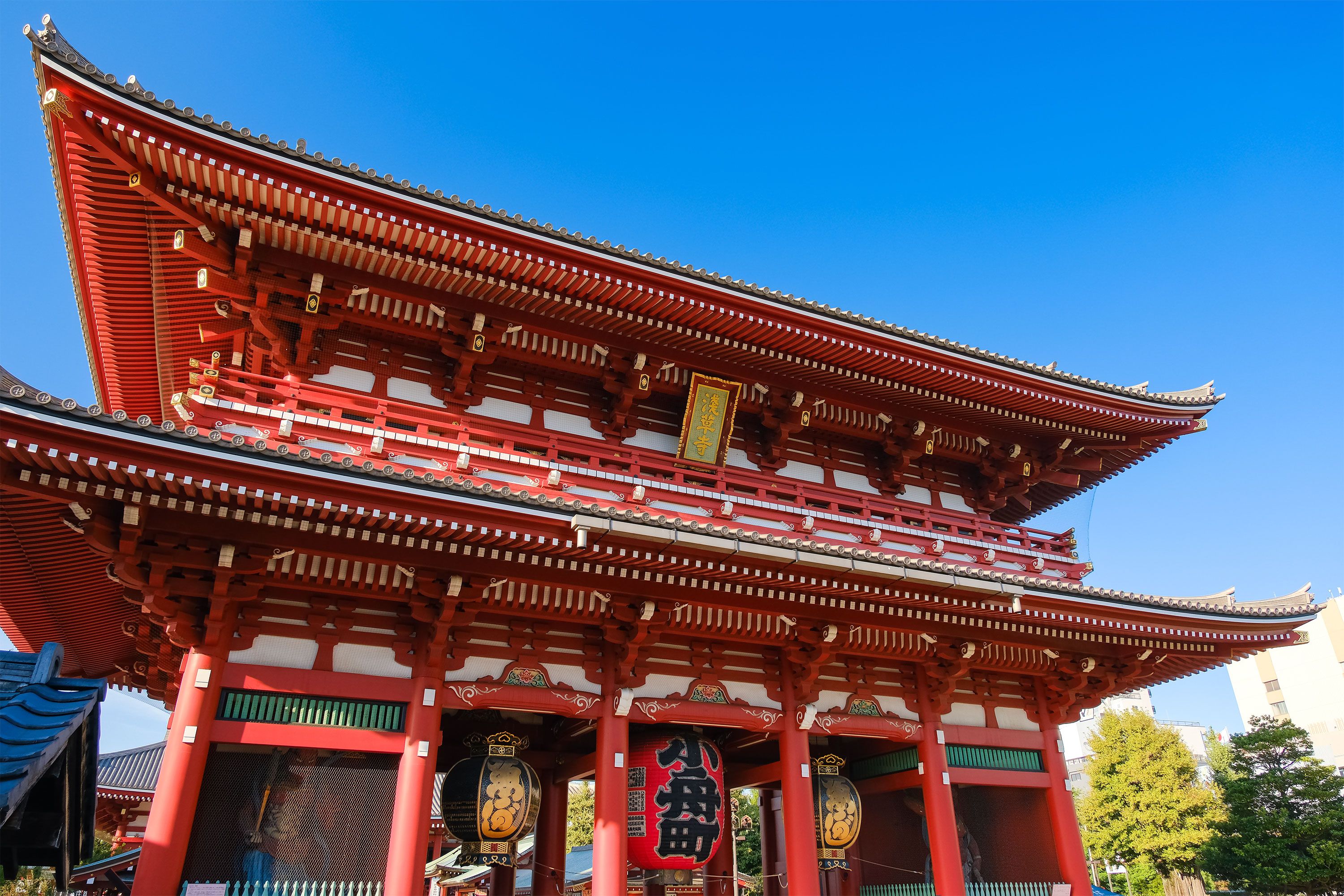A front view of a temple gate with ornate roof details, large lanterns, and temple visitors passing underneath.