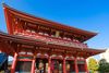 A front view of a temple gate with ornate roof details, large lanterns, and temple visitors passing underneath.