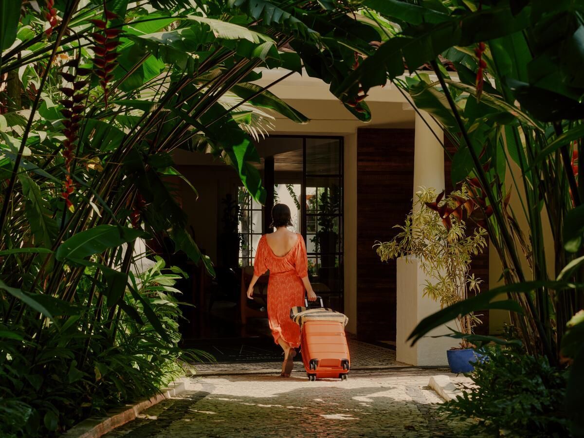 A woman in an orange dress wheeling an orange suitcase arrives at a hotel, through a tunnel of palms