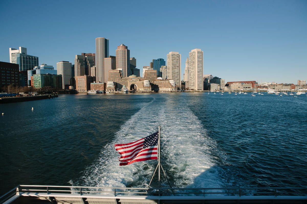 The US flag flies on the back of a boat sailing through Boston Harbor in Massachusetts on a sunny day