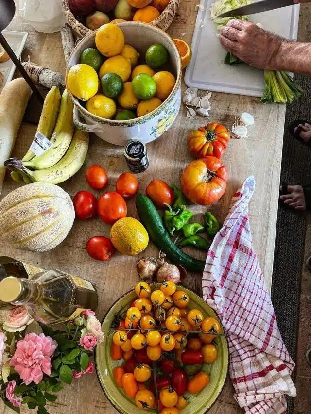 Overflowing bowls of tomatoes, lemons and limes share a busy kitchen table with fruit, as a person chops veg
