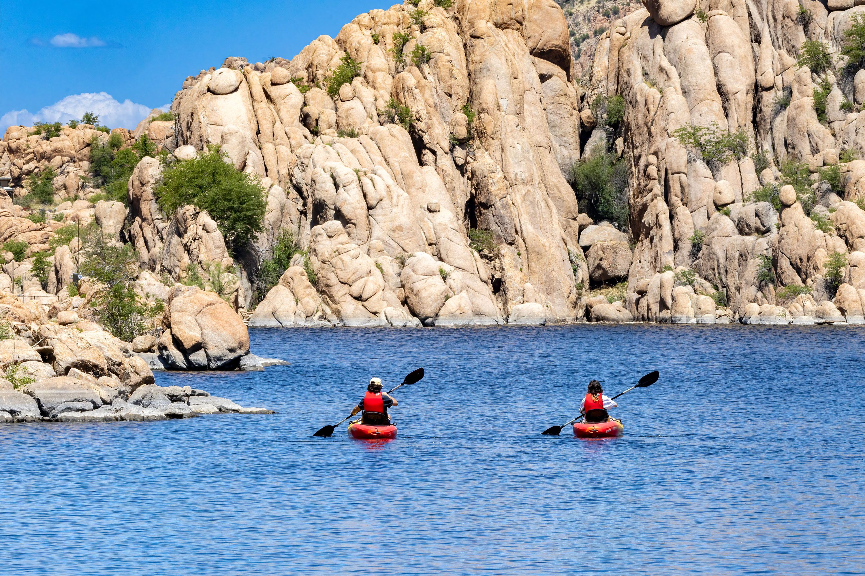 Two kayakers paddle across a calm lake beneath rugged granite cliffs.