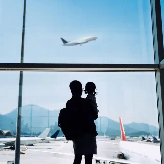 A father holds a child in front of an airport window, watching a plane taking off