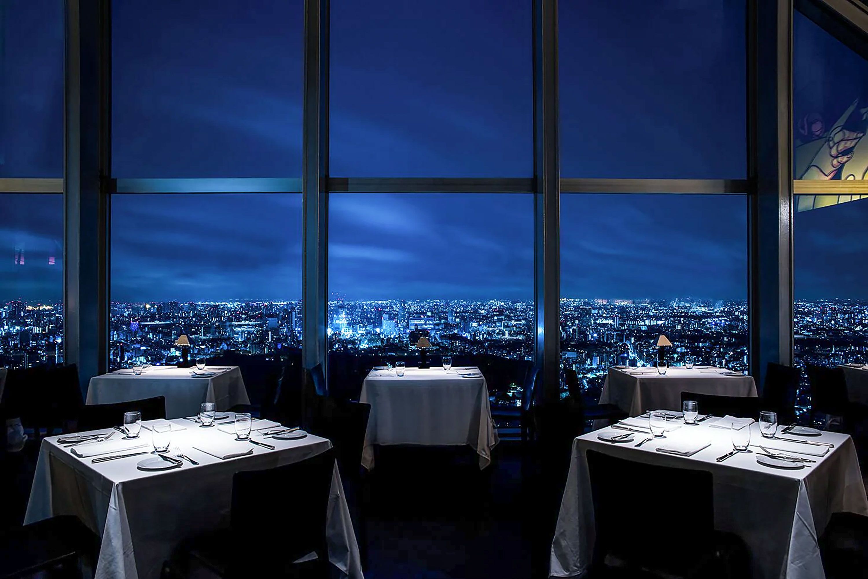 A dark diningroom with floor-to-ceiling windows and illuminated tables with view over city at night.