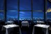 A dark diningroom with floor-to-ceiling windows and illuminated tables with view over city at night.