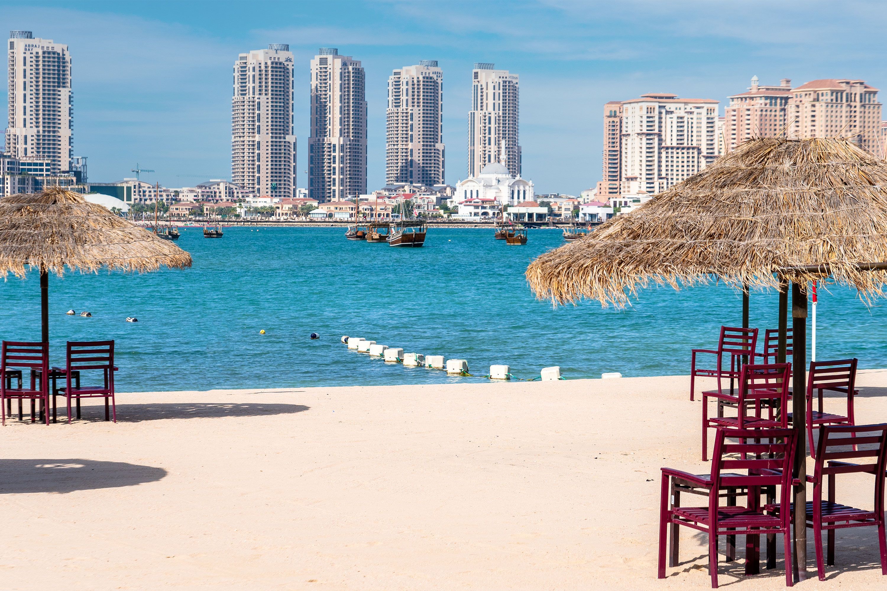 Tables and chairs arranged on the sandy shore of Katara Beach in Doha, overlooking the calm blue waters of the Arabian Gulf under a clear sky.