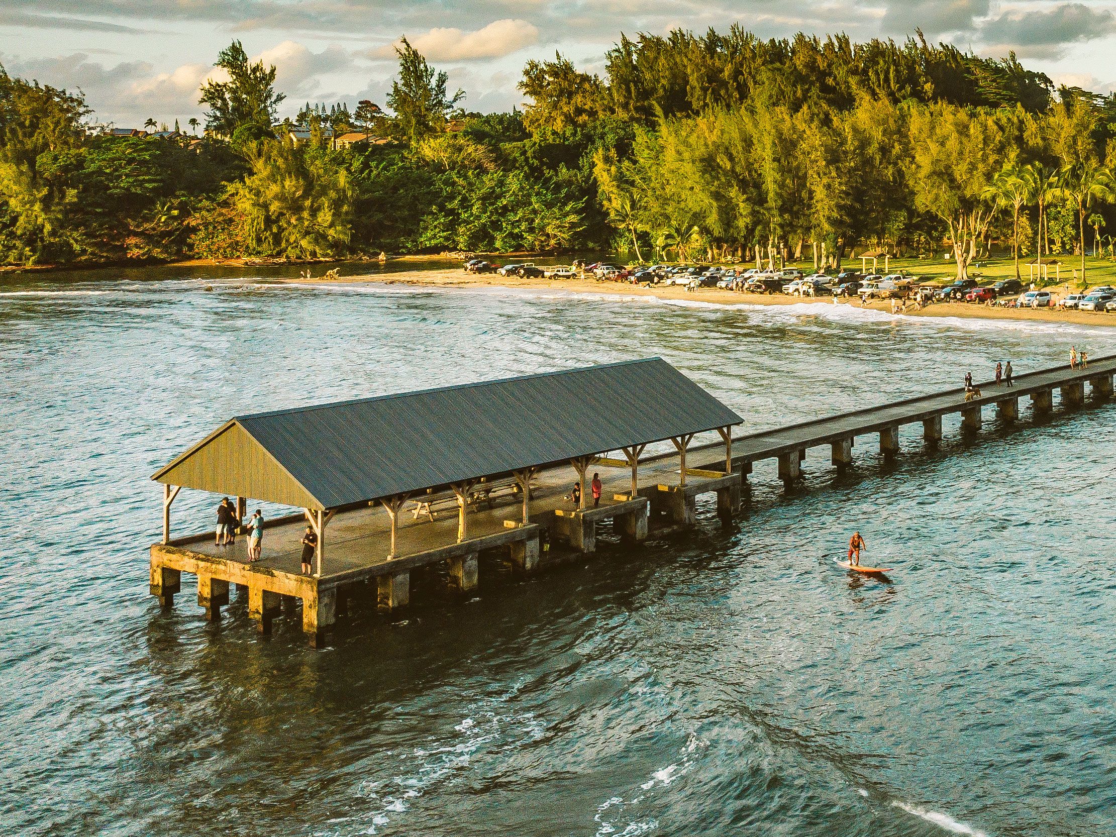 Ariel view from the water of people on the covered Hanalei pier and a person on a paddle board in the bay at sunset.
