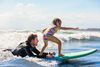 Little girl surfs while instructor in wet suit hangs on back of board.