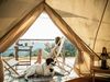 A view looking out of a canvas tent with a woman sitting in a folding chair reading a book on a small deck with her dog.