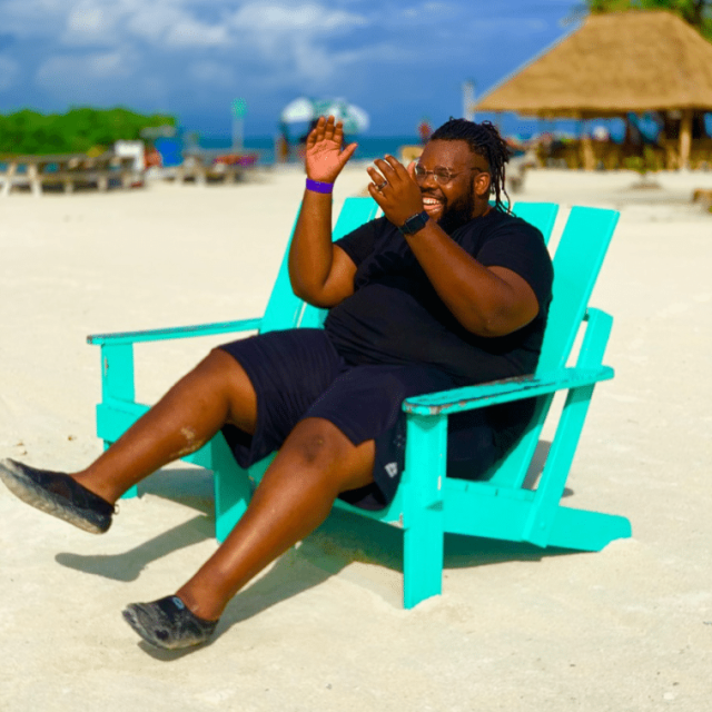 Travel influencer Jeff Jenkins sits on a beach in a bright turquoise wooden deckchair, laughing and clapping his hands