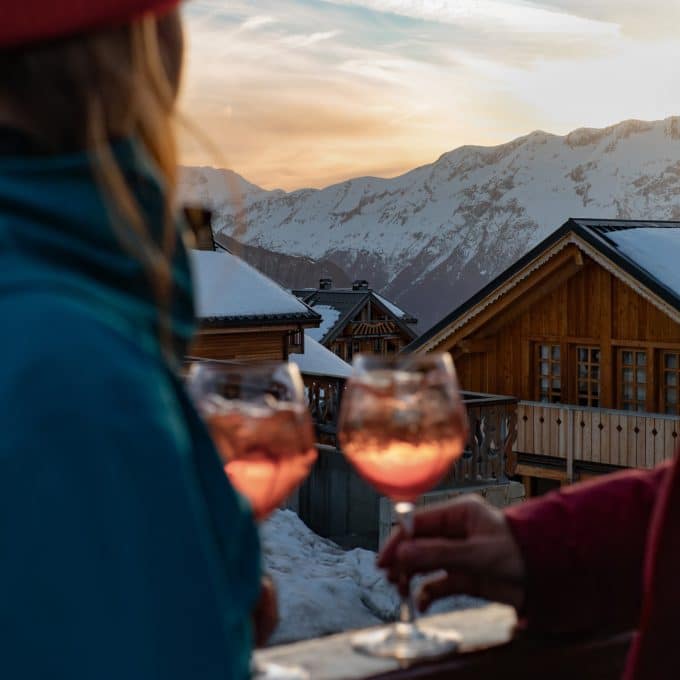 Two people drink Aperol spritzes on the balcony of a chalet in the snow-covered French Alps