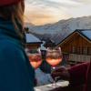 Two people drink Aperol spritzes on the balcony of a chalet in the snow-covered French Alps