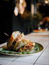 A sandwich sits on a decorative plate on a white-tiled counter in a restaurant
