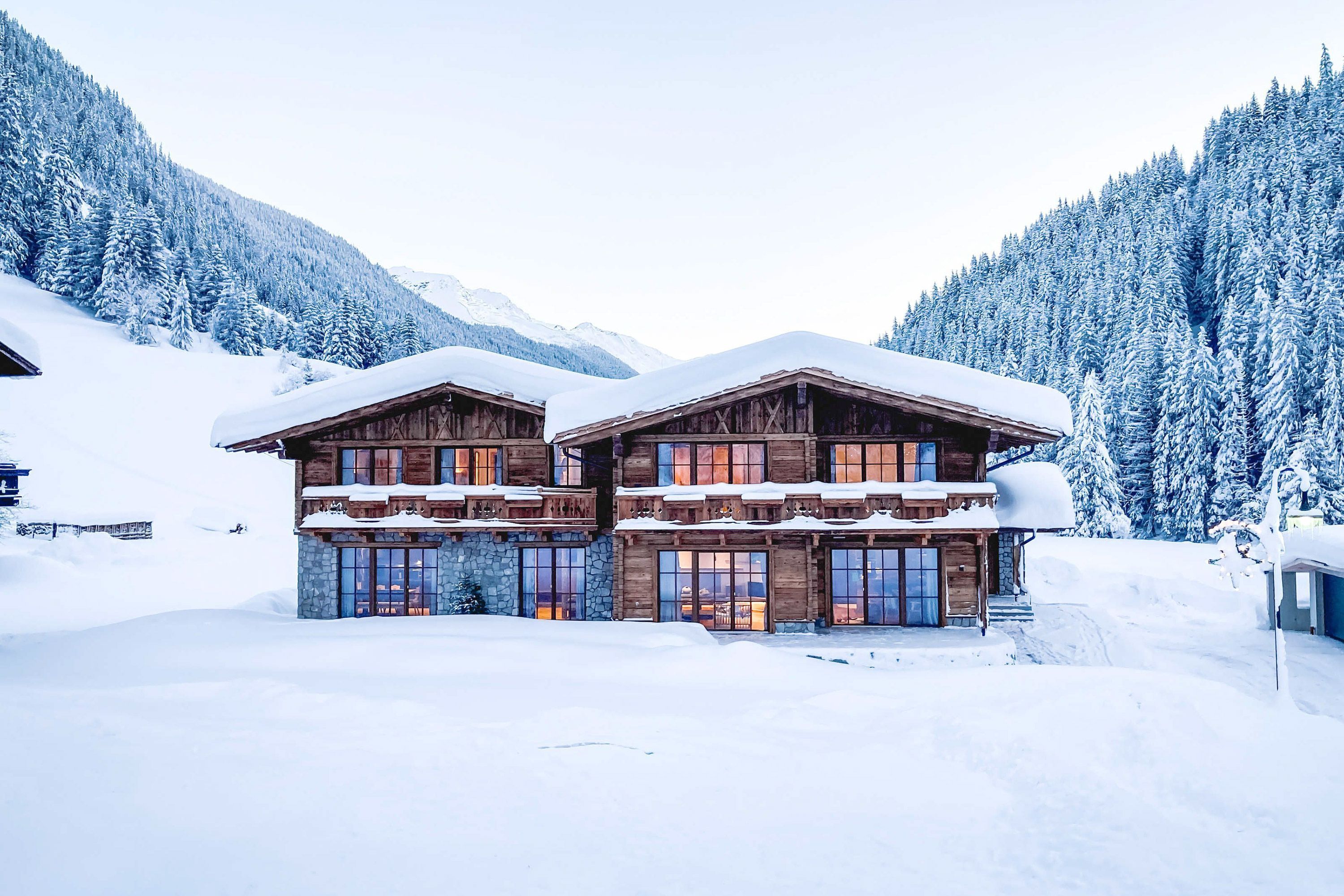 Ein Doppelhaus in alpiner Holzbauweise mit einer dicken Schneeschicht auf dem Dach steht in einer verschneiten Berglandschaft 
