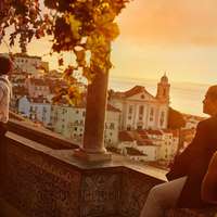 People sit on the wall, decorated with prettily decorated tiles, at the Miradouro de Santa Luzia, and gaze at Lisbon's white-washed, red roofed building below