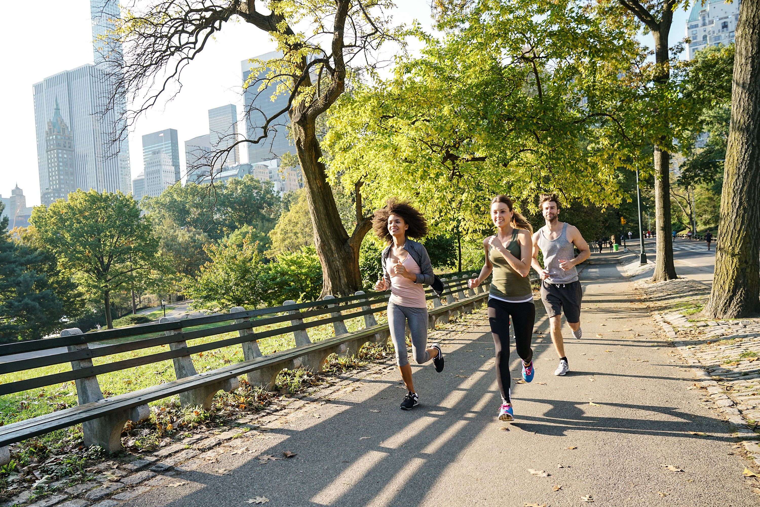 Three runners jog through park on asphalt path lined with benches surrounded by grass, trees, and skyscrapers in the background. 