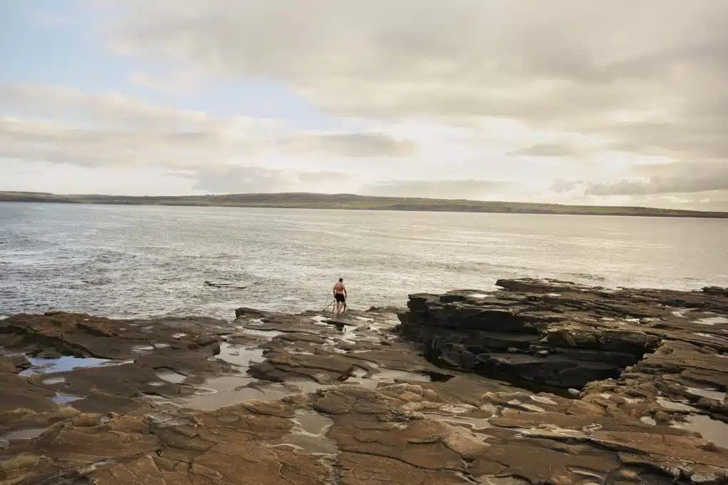 A swimmer looks out over the water from the rocks as sun breaks through the clouds at secluded Clahane Beach
