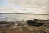 A swimmer looks out over the water from the rocks as sun breaks through the clouds at secluded Clahane Beach