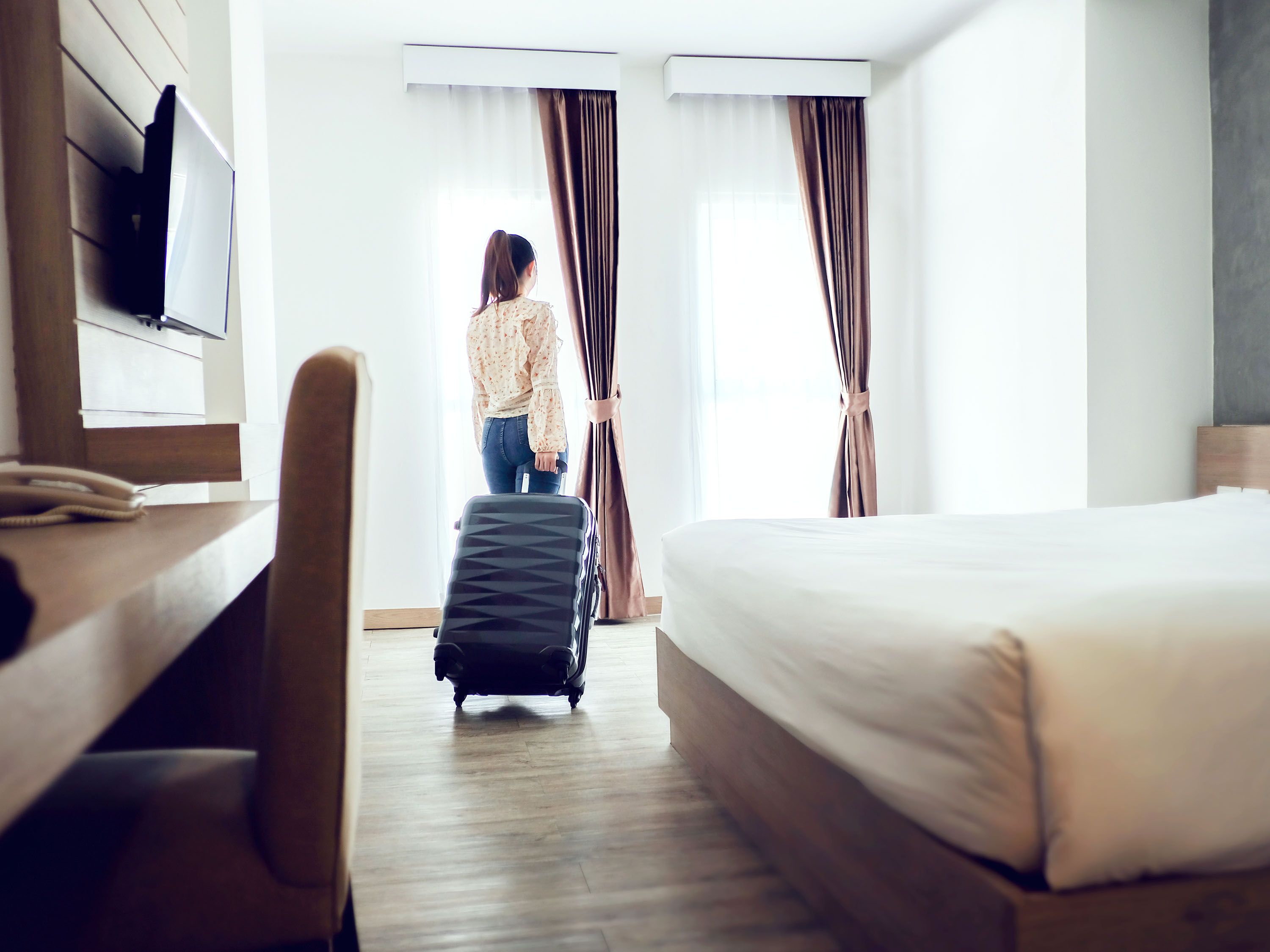 Back of woman dragging suitcase into crisp hotel room.