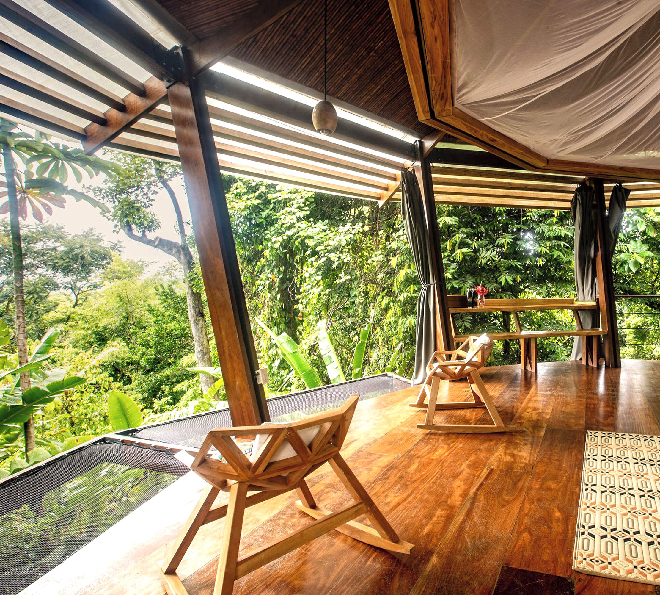 Open air hotel room with natural wood chairs overlooking green forest area.