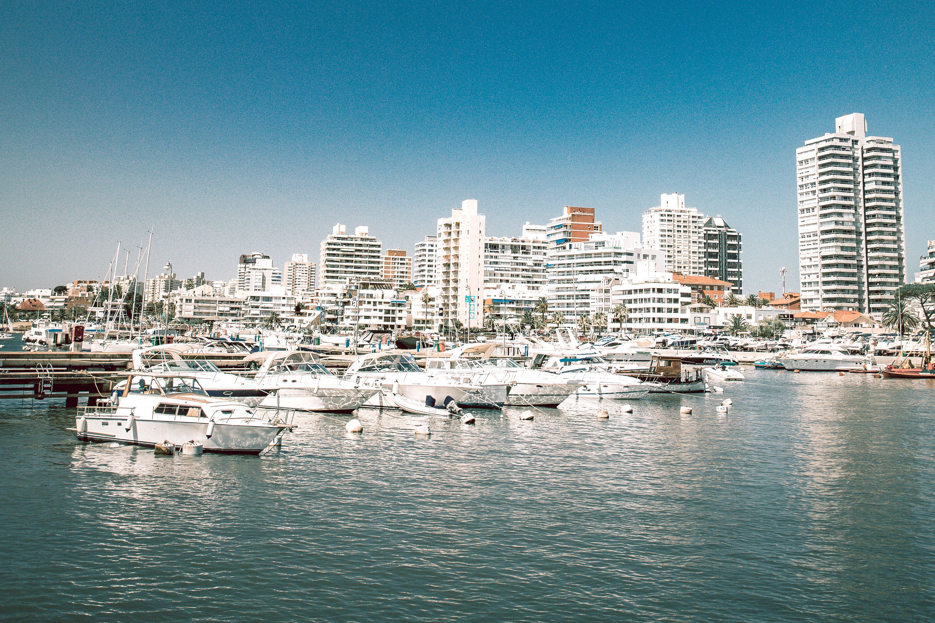 Pleasure boats are moored in a harbor surrounded by city buildings.