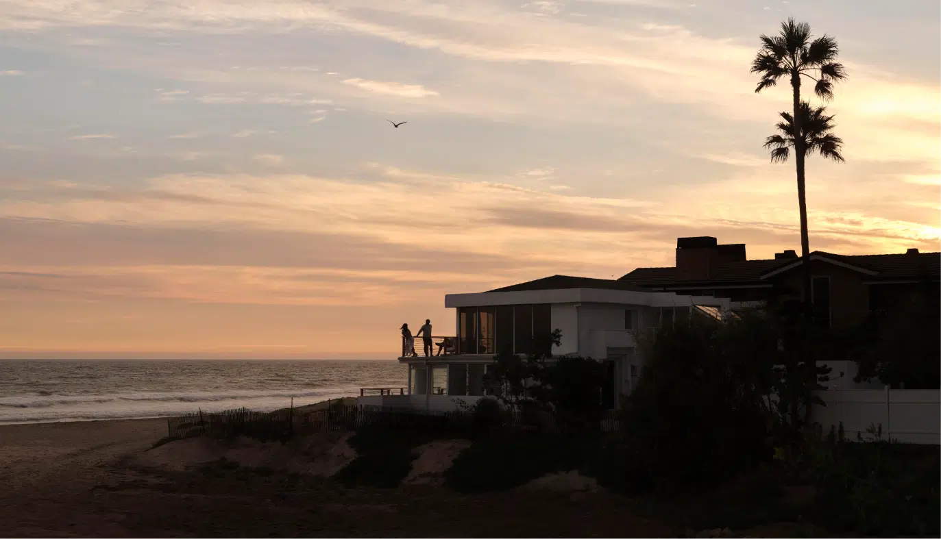 Seen from a distance, a couple stand on the wraparound balcony of a beach front home, as the sun sets