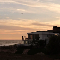 Seen from a distance, a couple stand on the wraparound balcony of a beach front home, as the sun sets