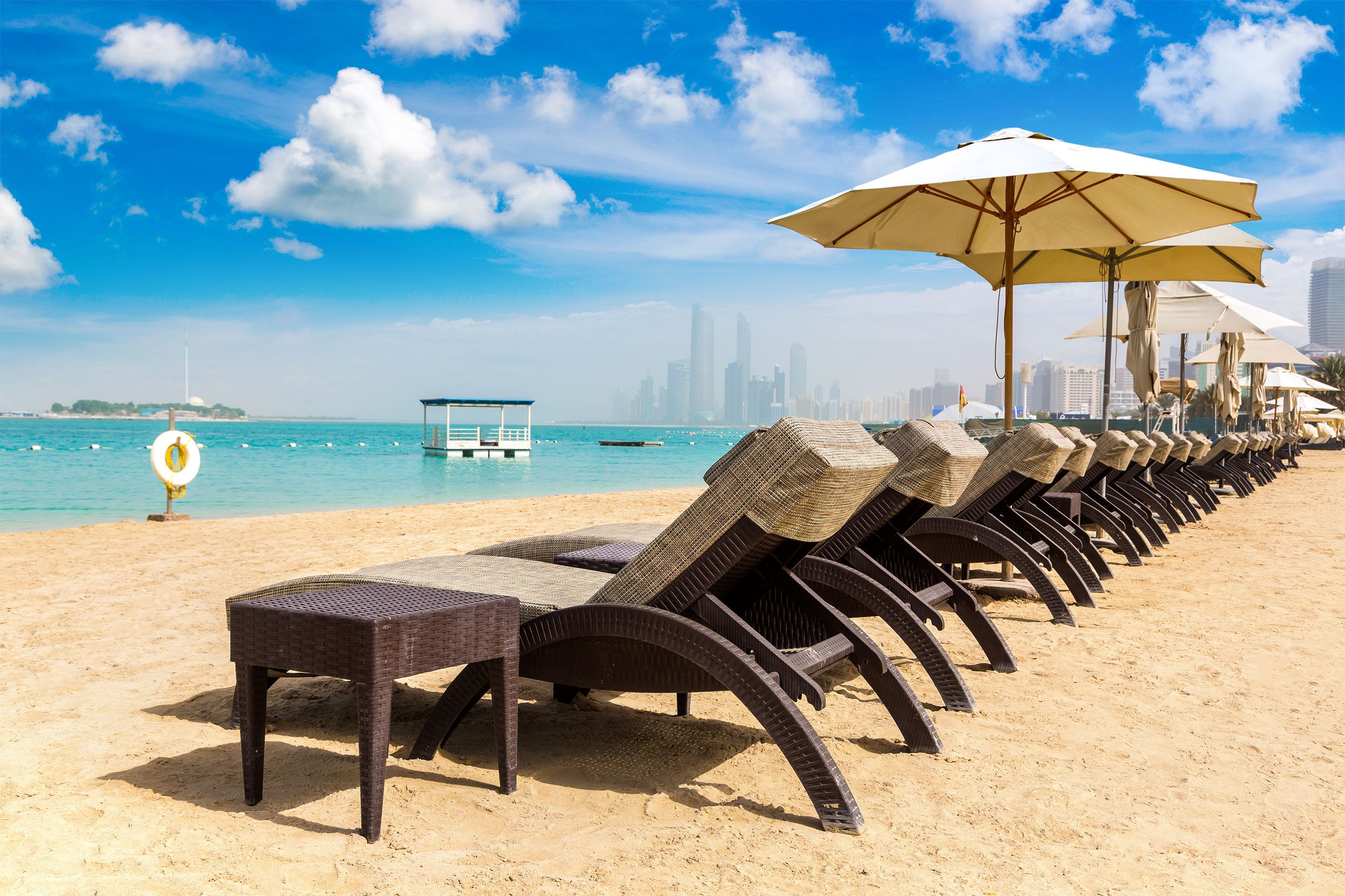 Rows of empty sunbeds and straw parasols set on a sandy Abu Dhabi shoreline, with the turquoise gulf water and a clear sky in the background.