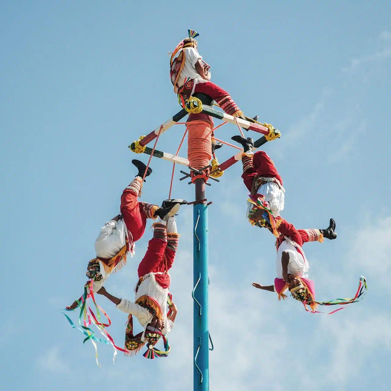 Dressed in red and white with streamers, a group of people hang upside down from a colorful pole to perform the Mesoamerican ritual of the voladores 