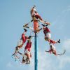 Dressed in red and white with streamers, a group of people hang upside down from a colorful pole to perform the Mesoamerican ritual of the voladores