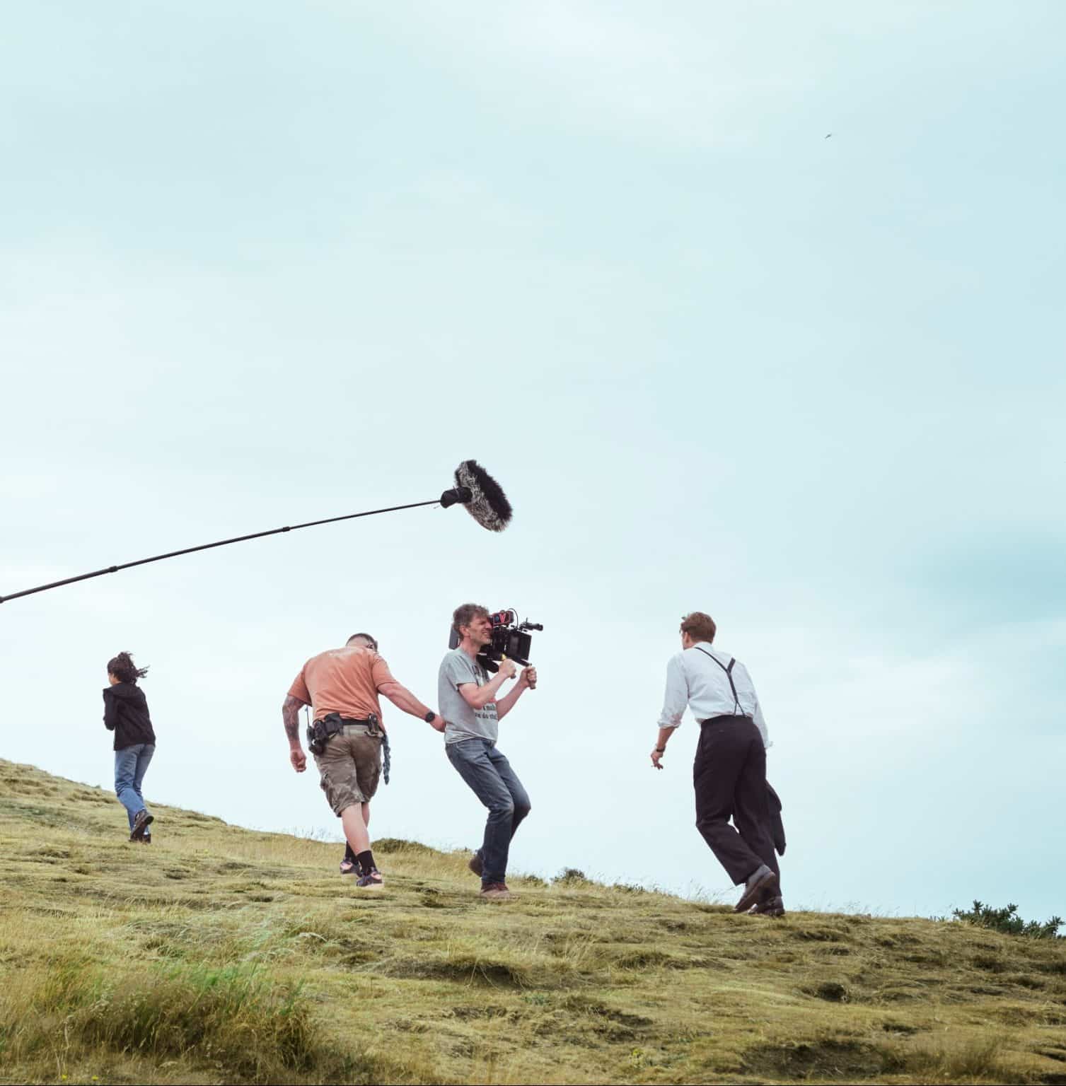With a camera crew and microphone boom in front of him, actor Leo Woodall climbs Arthur's Seat in Edinburgh, in a behind-the-scenes shot from One Day (photo courtesy of Netflix)