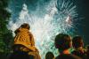 A child on a parent's shoulders stands among a crowd of people watching fireworks explode overhead